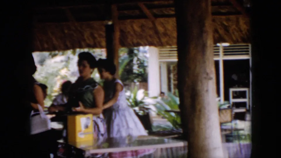 1956 Cuba frame showing people at a shaded roadside or rest area beneath a thatched roof after the bus sequence.