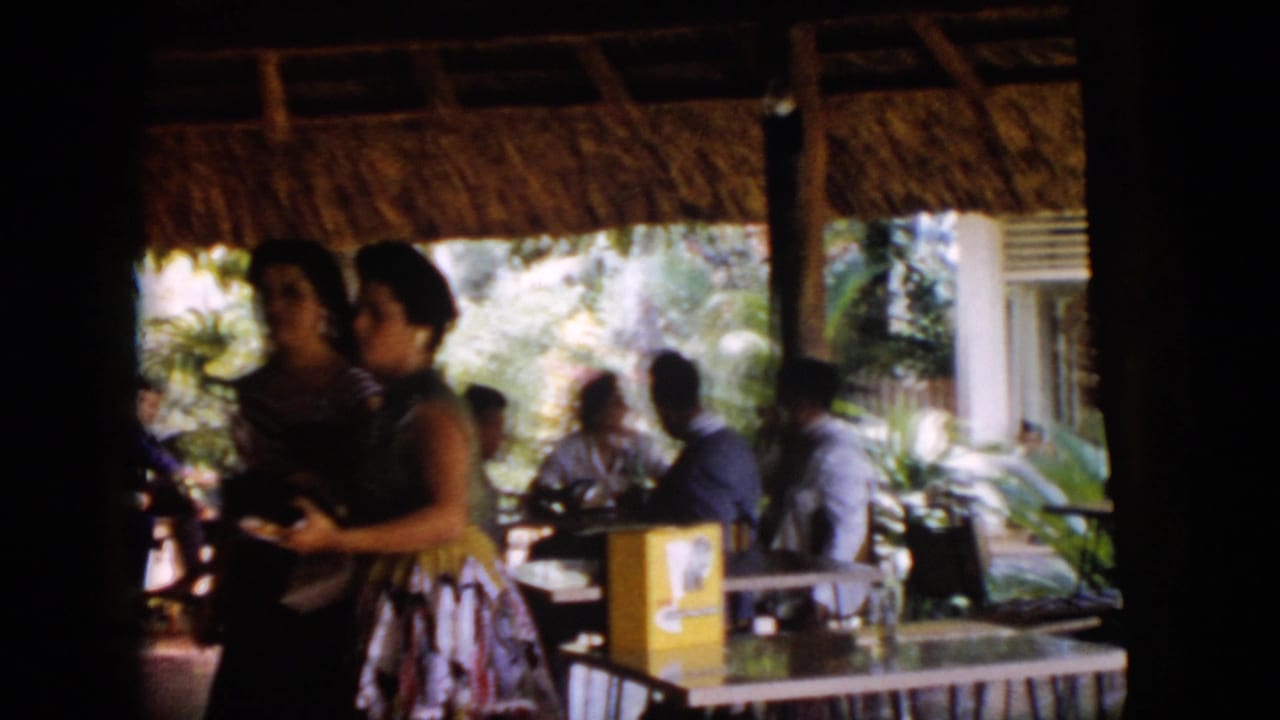 1956 Cuba home-movie frame showing guests in mid-century attire under a thatched open-air pavilion with dining tables nearby.