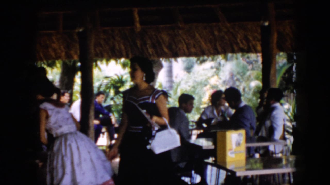 1956 Cuba frame with women in formal daytime dresses passing seated guests inside a tropical thatched pavilion.