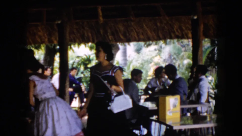 1956 Cuba frame with women in formal daytime dresses passing seated guests inside a tropical thatched pavilion.