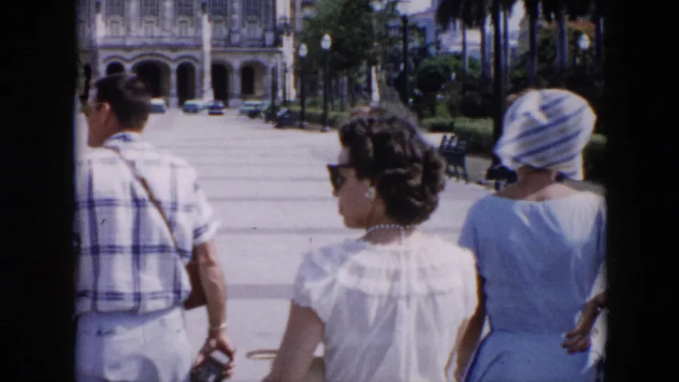 1956 Cuba street scene with tourists in light clothing walking past palms and an ornate arched colonial-style building.