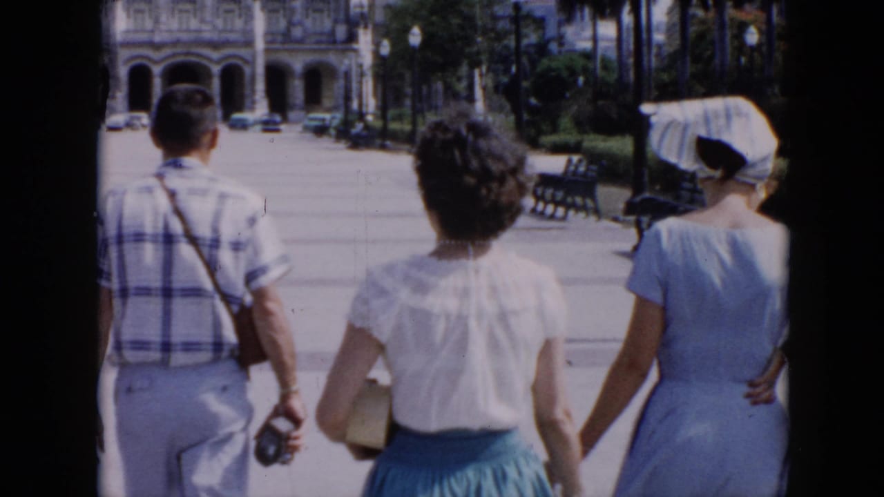 In 1956 Cuba home-movie footage, three adults walk away from the camera through a sunny park promenade with benches and palms.