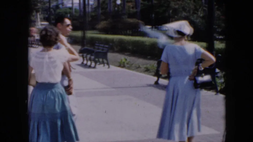 The handheld camera follows the adults from behind as they pass benches, planted beds, and palms in a sunny Cuban park setting.