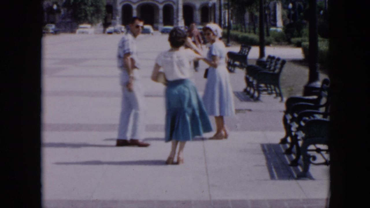 Three adults stand on a wide sidewalk near benches and palm trees.