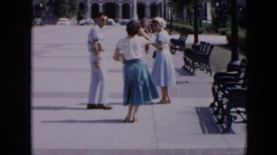 1956 Cuba sidewalk scene with a man and two women standing near benches, with palm trees and arched buildings in the background.