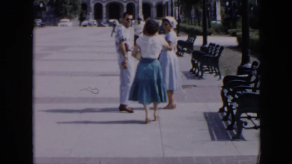 1956 Cuba home-movie frame showing three adults on a broad public sidewalk, with benches and arched architecture behind them.
