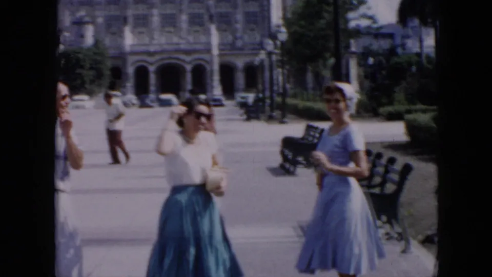 1956 Cuba frame with two women in mid-century dresses smiling or posing near benches as a man walks in the background.