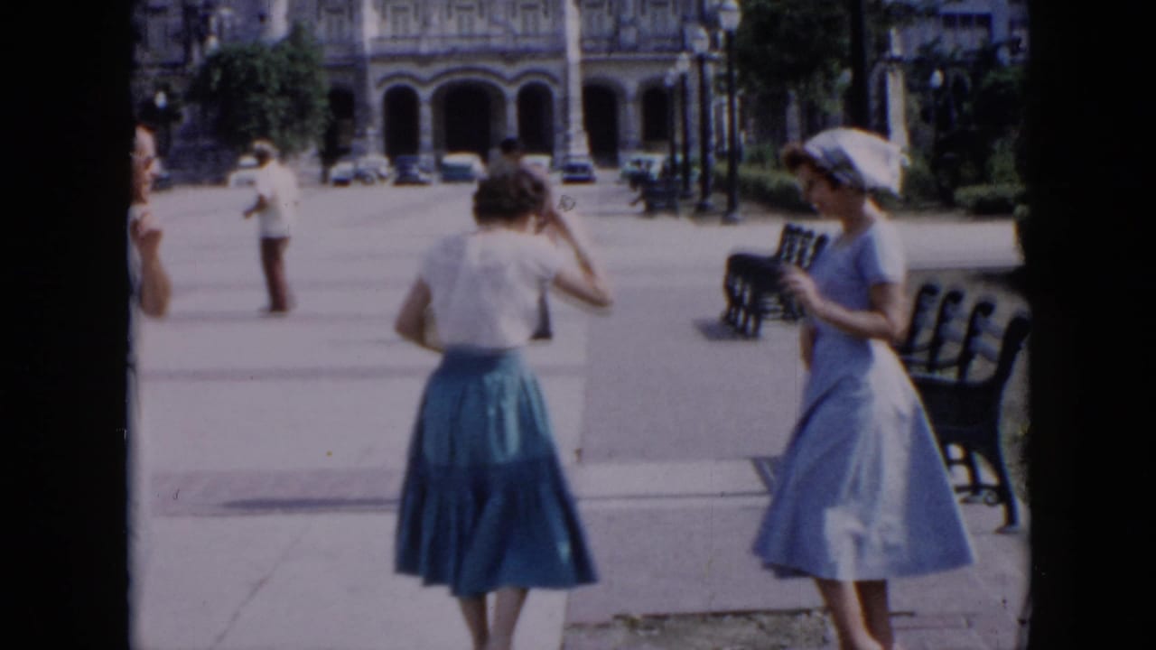 In 1956 Cuba home-movie footage, two women in mid-century dress appear in a plaza with benches, pedestrians, and an arched building behind them.
