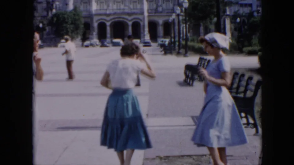 In 1956 Cuba home-movie footage, two women in mid-century dress appear in a plaza with benches, pedestrians, and an arched building behind them.