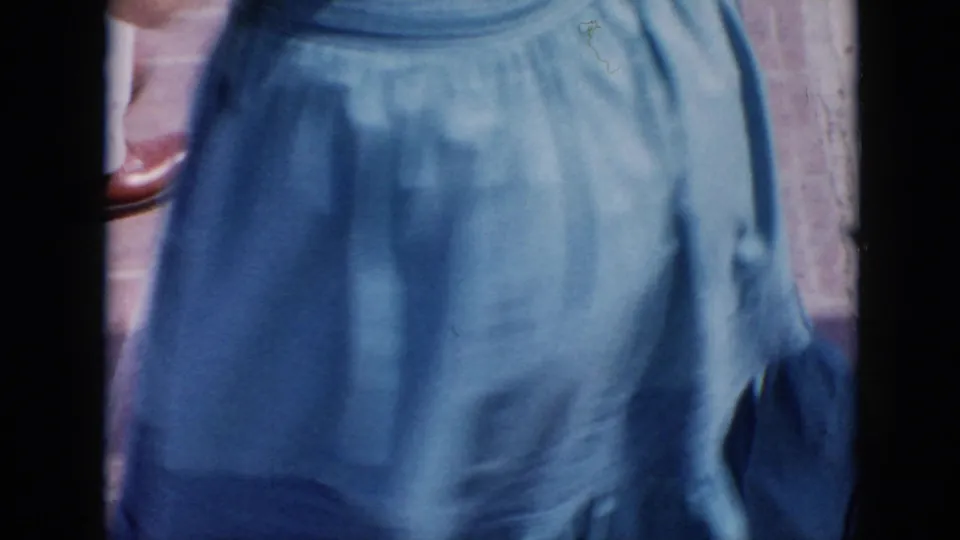 A close, cropped 8mm frame shows the blue skirt of one woman as the handheld camera passes very near her.