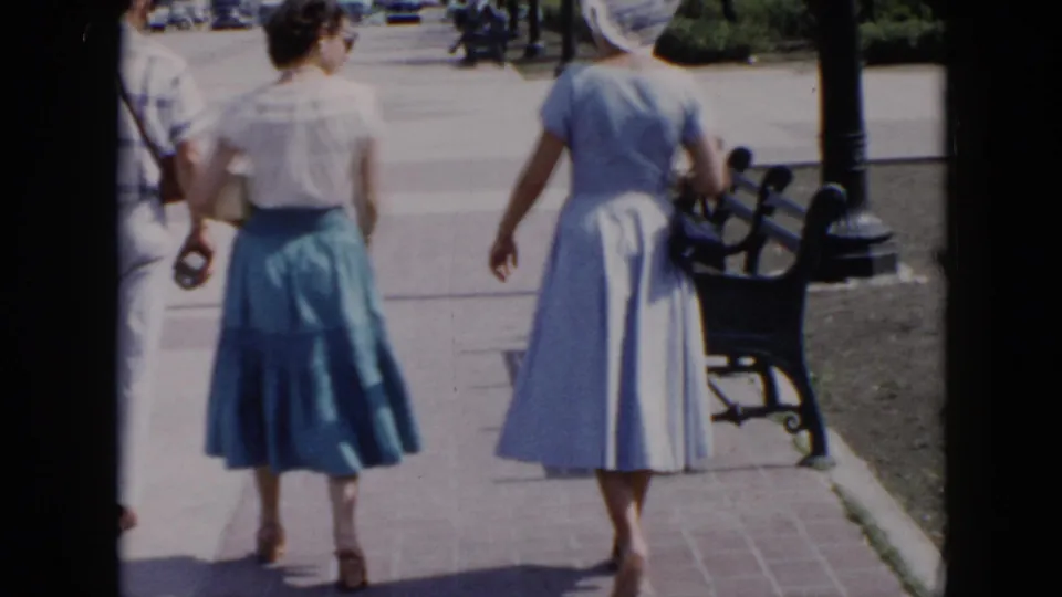 Two women in 1950s clothing walk away through a Cuba plaza, with a black bench, lamppost, pavement, and partial greenery visible.