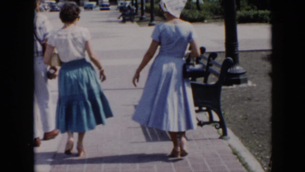 In 1956 Cuba footage, three adults walk away on a paved park path with benches, lampposts, lawn, and distant cars visible.