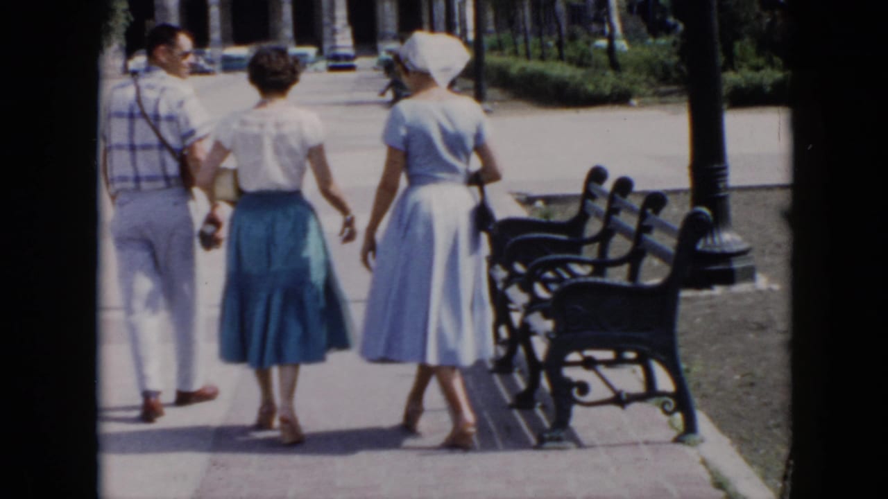 A man and two women walk away beside benches and a lamppost in a park.