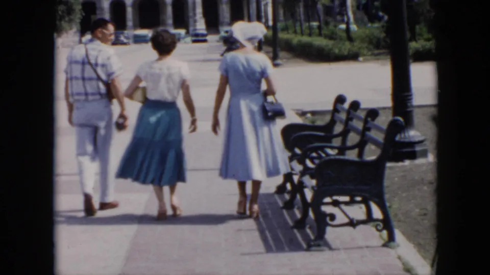 In the 1956 Cuba home movie, three adults move farther along the path, with black benches, lawn, trees, and arched architecture ahead.
