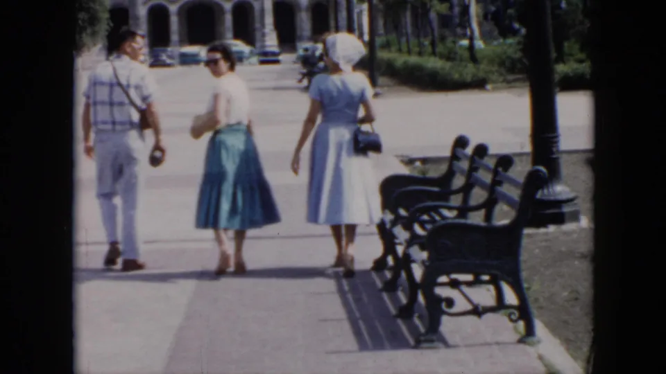 A woman in a blue skirt turns partly toward the camera as the group walks through the Cuban park, with benches and arched buildings nearby.