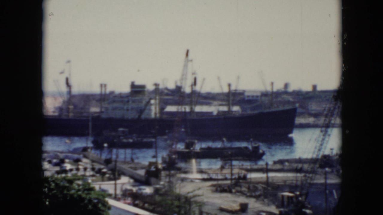 1956 Cuba home-movie frame showing a large ship in a harbor, viewed through a window with cranes, dockside roads, and trees in the foreground.
