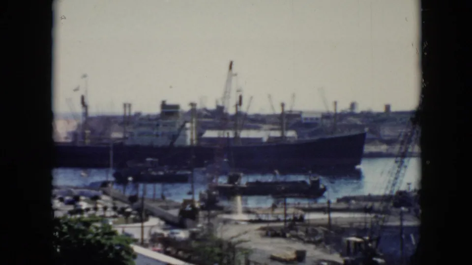 1956 Cuba home-movie frame showing a large ship in a harbor, viewed through a window with cranes, dockside roads, and trees in the foreground.