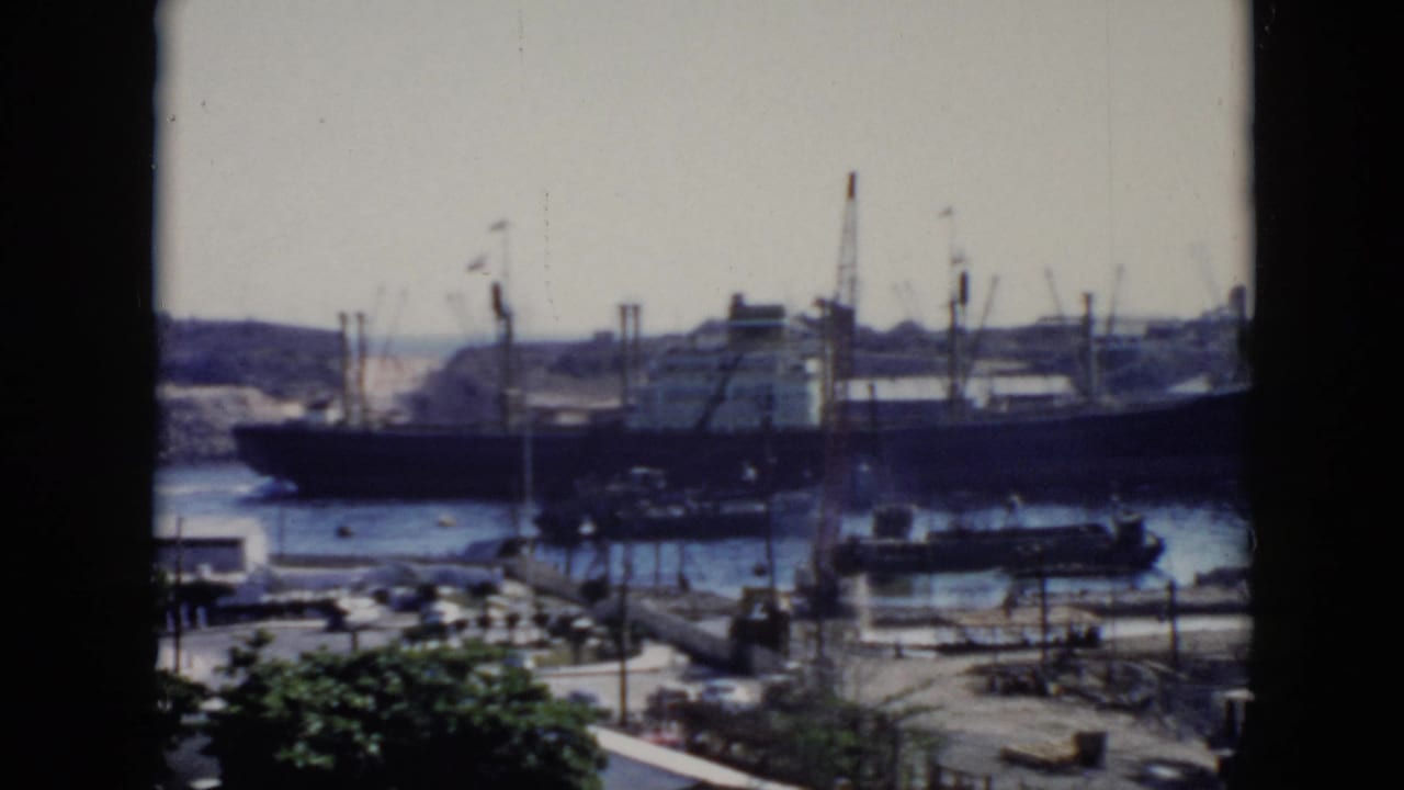 Large harbor ship near cranes and smaller boats, framed by a dark vertical window divider.