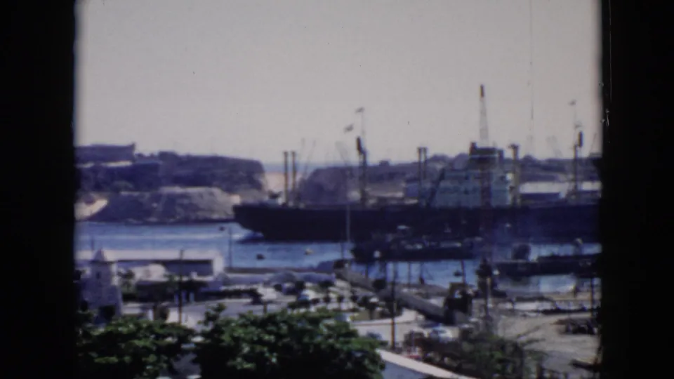 Vintage 8mm Cuba harbor frame showing a large ship partly behind a window edge, with cranes, shoreline structures, and foreground trees.