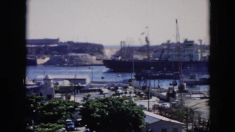 1956 Cuba home-movie view of a large ship near the right side of the harbor, with cranes, small vessels, dock roads, and trees visible.