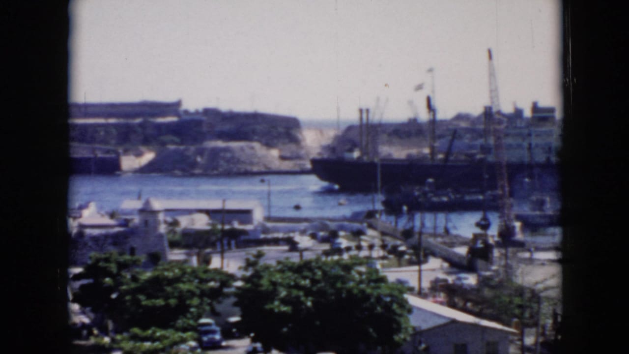1956 Cuba harbor scene seen through a dark window frame, with docked ships, cranes, blue water, white buildings, trees, and street traffic below.