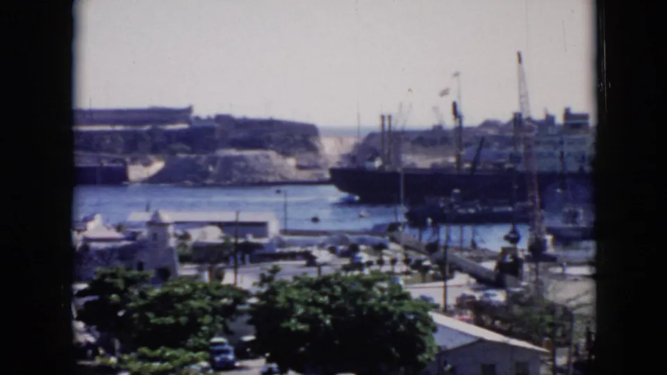 1956 Cuba harbor scene seen through a dark window frame, with docked ships, cranes, blue water, white buildings, trees, and street traffic below.