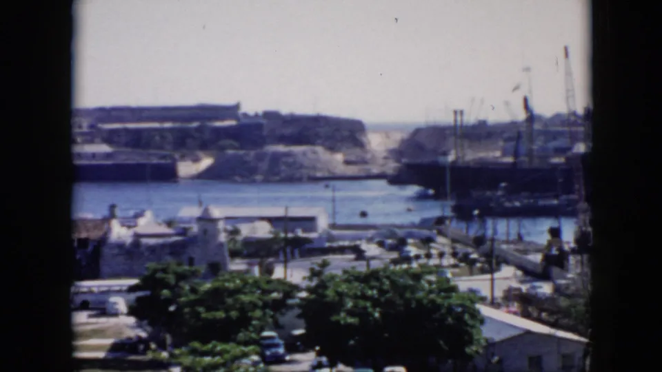 Cuba harbor view from 1956 with a window mullion in front, showing moored vessels, industrial cranes, a stone structure, low buildings, trees, and cars.