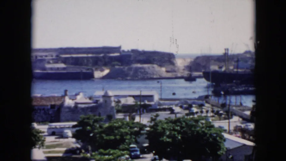Soft 8mm frame of a 1956 Cuba waterfront, showing harbor water, docked ships, cranes, a masonry structure, white buildings, trees, and cars below.