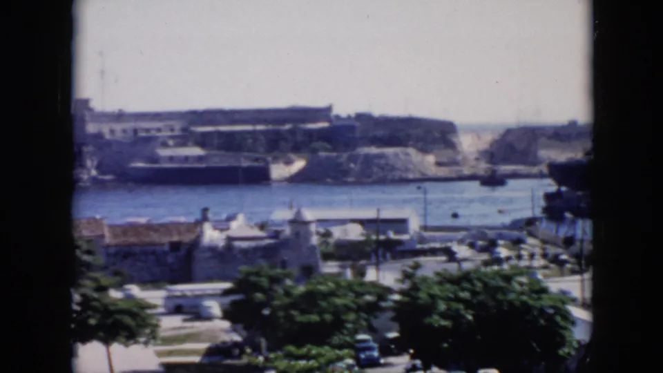 Final 1956 Cuba harbor frame with blue water, ships and cranes beyond a waterfront road, white buildings, trees, parked cars, and dark film-frame edges.