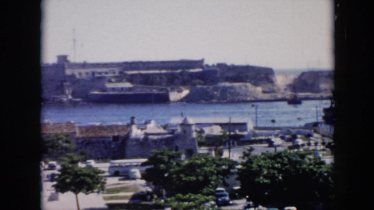 1956 Cuba 8mm view across a harbor toward a stone coastal fortress, with trees, cars, and rooftops in the foreground.