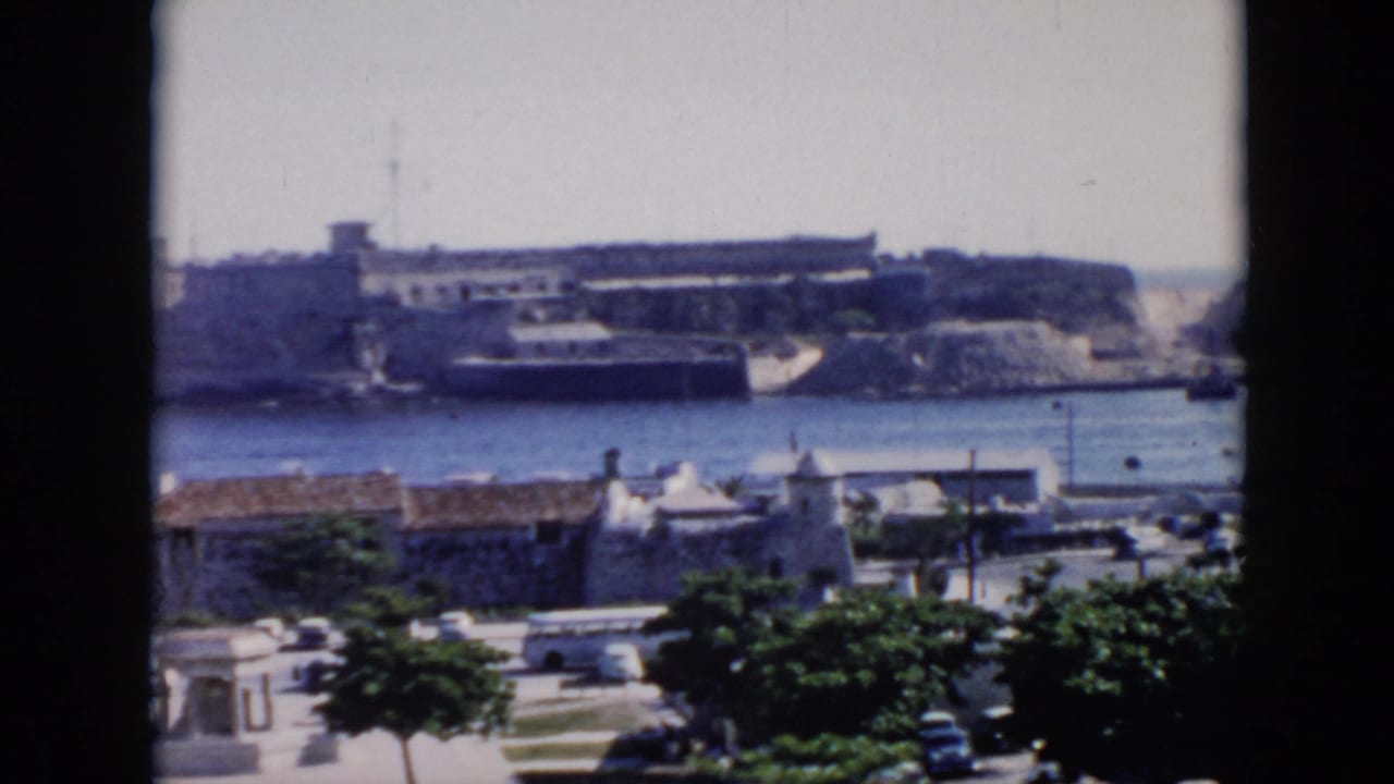 1956 Cuba home-movie frame showing a coastal fortification across calm water, with mid-century traffic and greenery in front.