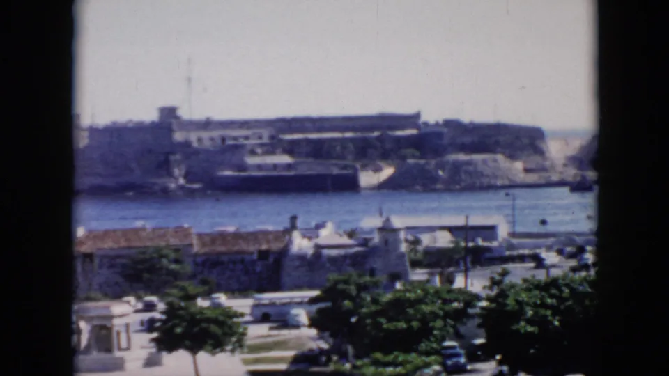 1956 Cuba home-movie frame showing a coastal fortification across calm water, with mid-century traffic and greenery in front.