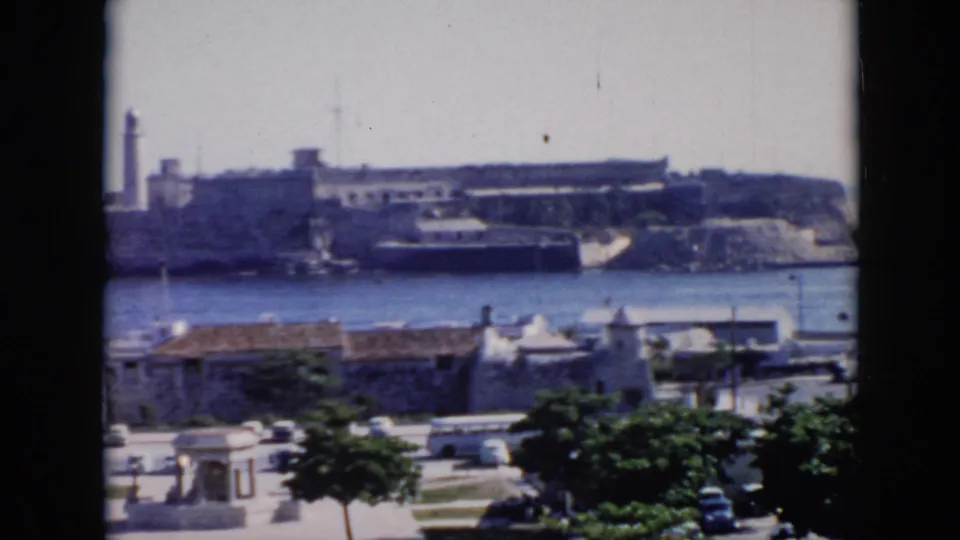 1956 Cuba 8mm frame with a white bus, parked cars, foreground greenery, and a fortress-like shoreline across the harbor.