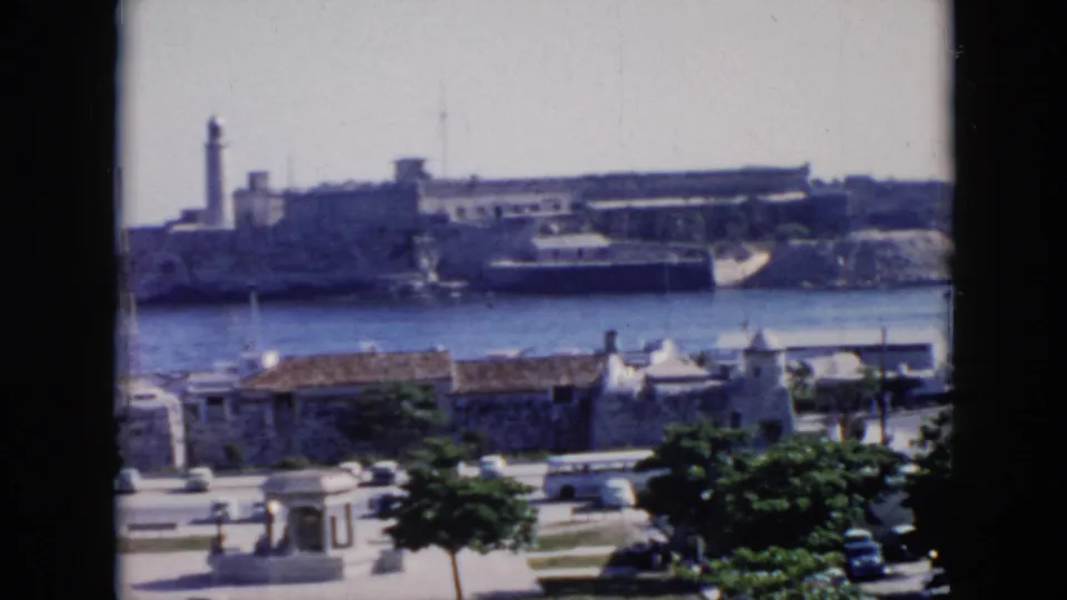 1956 Cuba harbor overlook framed by dark film edges, showing stone coastal buildings, a tower, cars, trees, and a white bus.