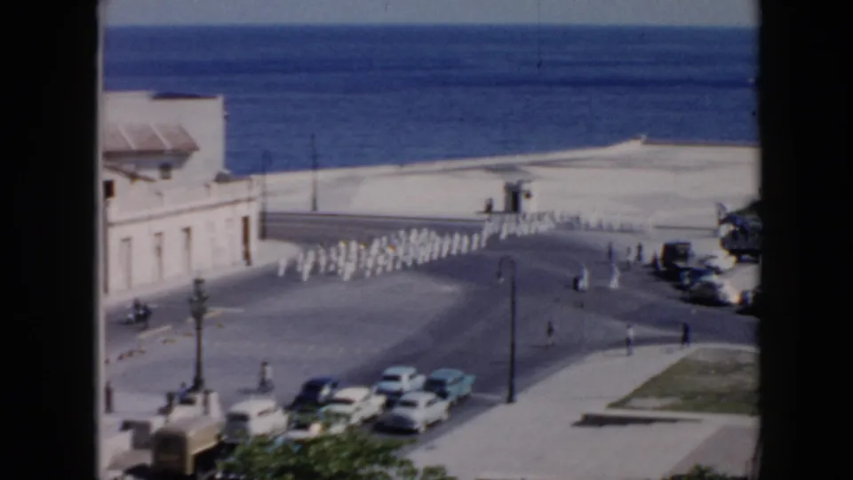 1956 Cuba home-movie frame looking down on a coastal street as uniformed personnel march near pedestrians, parked cars, and a seawall.