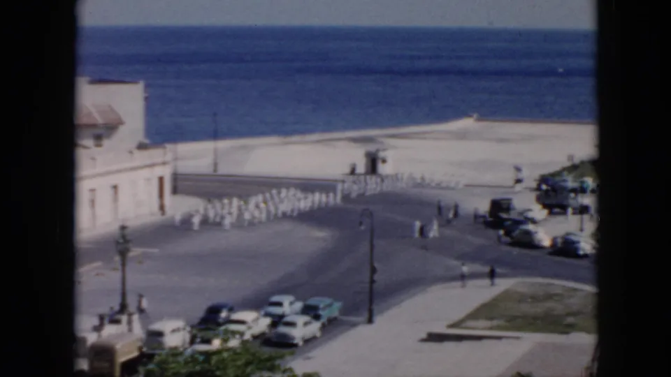 1956 Cuba waterfront frame showing a uniformed procession near the seawall, with pedestrians, parked cars, streetlamps, and open water in the background.