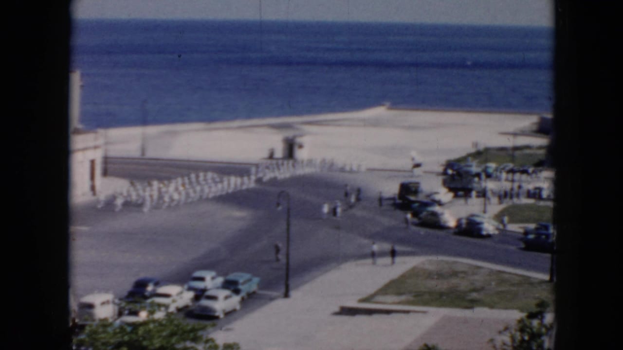 1956 Cuba home-movie frame showing soldiers in white uniforms marching near a waterfront road with parked cars and ocean beyond.