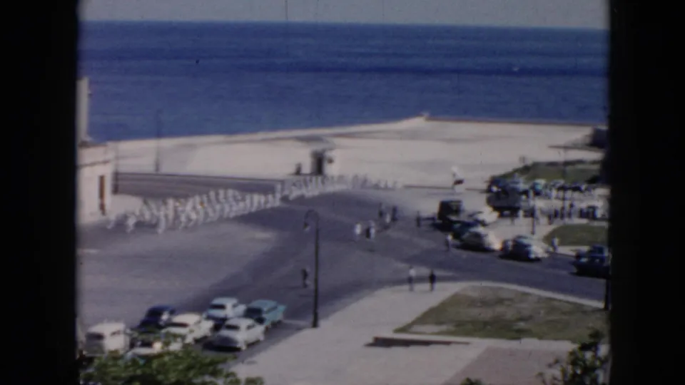 1956 Cuba home-movie frame showing soldiers in white uniforms marching near a waterfront road with parked cars and ocean beyond.