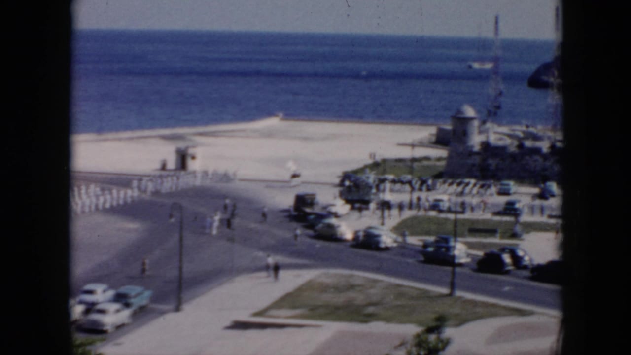 1956 Cuba frame with soldiers marching by a coastal street, mid-century cars, pedestrians, ocean water, and a small fort-like building.