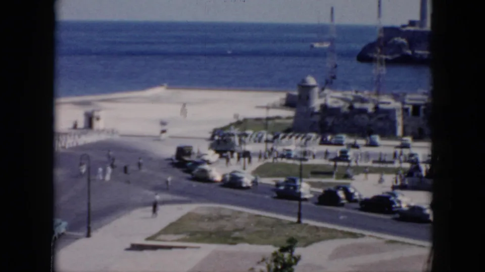 1956 Cuba home-movie view of a coastal parade area with soldiers, classic cars, pedestrians, ocean, and waterfront structures.
