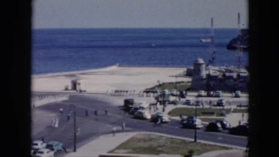 1956 Cuba frame showing a wide coastal road scene with soldiers in formation, parked cars, pedestrians, beach, and blue ocean.