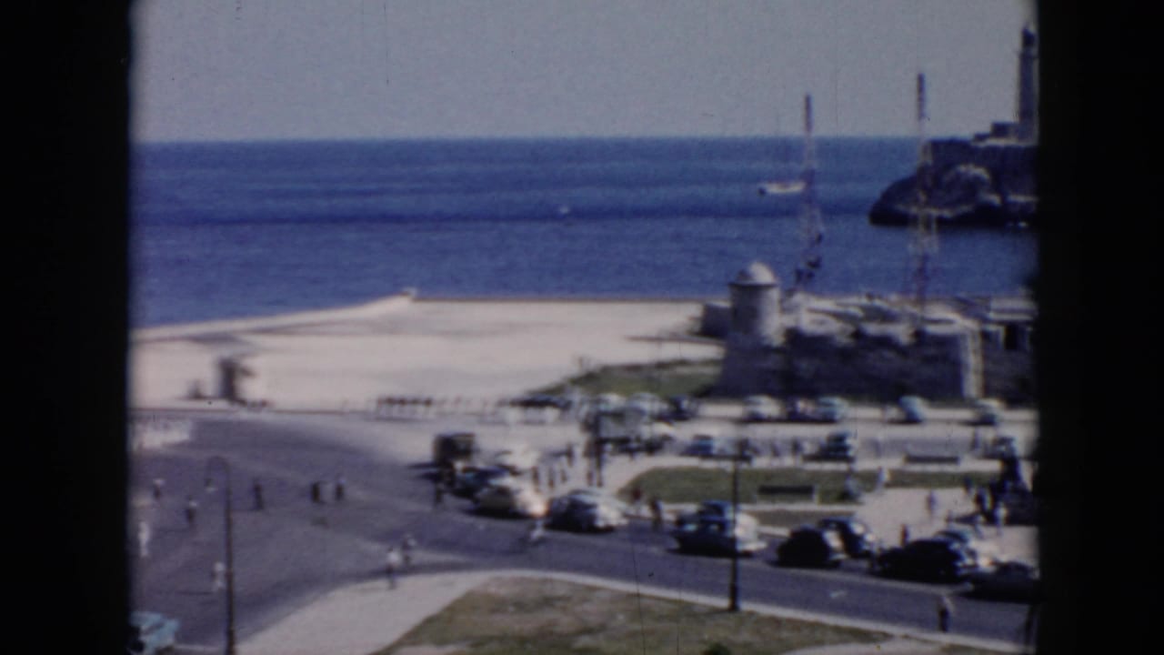 1956 Cuba 8mm frame showing a coastal road with mid-century cars, pedestrians, ocean, and a lighthouse near stone waterfront structures.