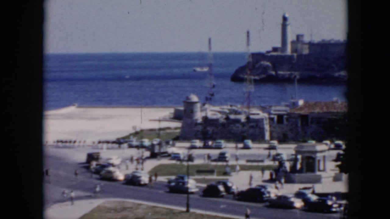1956 Cuba home-movie frame of traffic and pedestrians near a waterfront promenade, with ocean and a lighthouse on a rocky point.