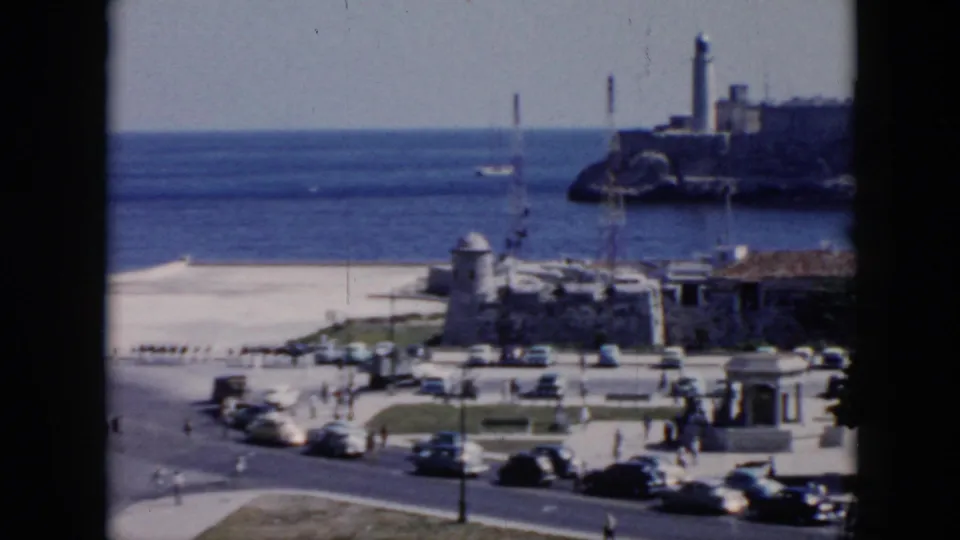 1956 Cuba home-movie frame of traffic and pedestrians near a waterfront promenade, with ocean and a lighthouse on a rocky point.
