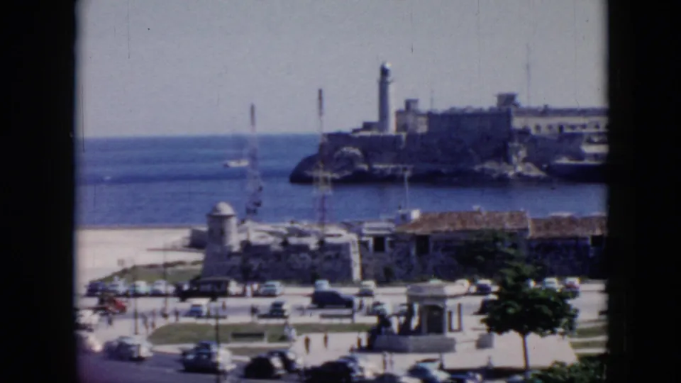1956 Cuba 8mm frame showing a harbor-side road and promenade below stone buildings, with cars, pedestrians, trees, and a lighthouse across the water.