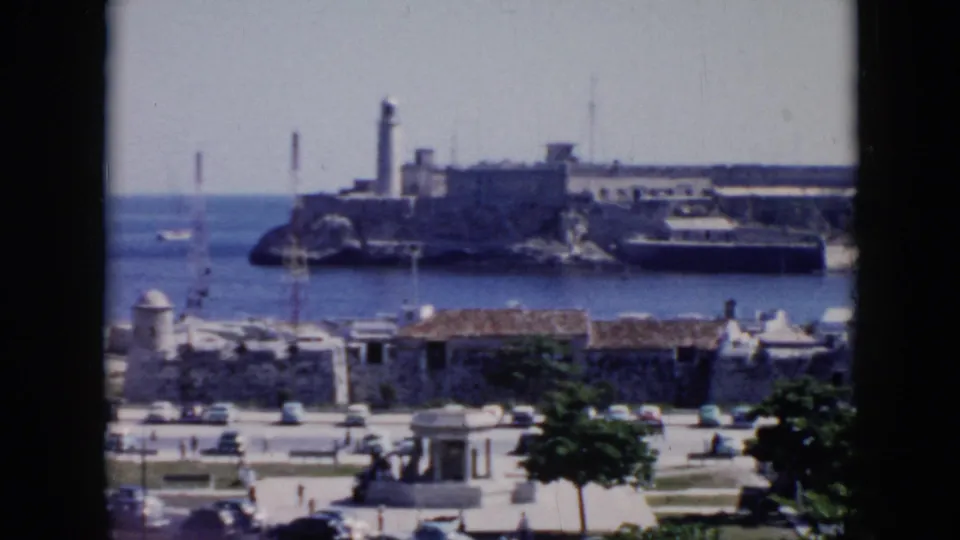1956 Cuba frame of a waterfront road with vintage cars and pedestrians, looking toward a lighthouse and fort-like stone complex across the harbor.