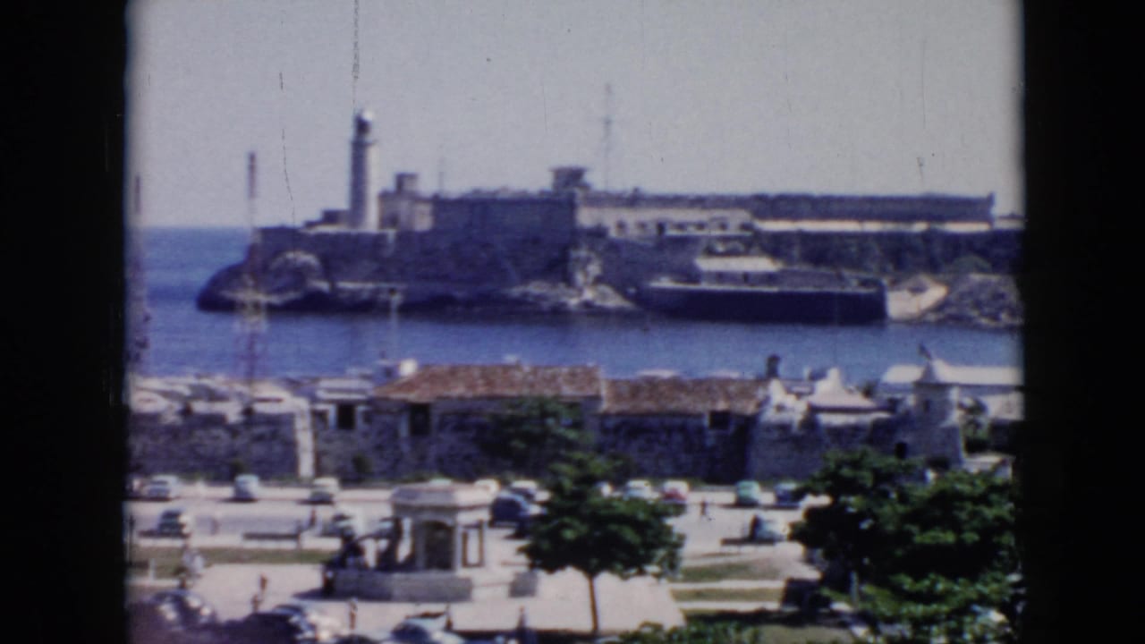 Coastal fortress with lighthouse across blue water, seen above waterfront buildings and trees.