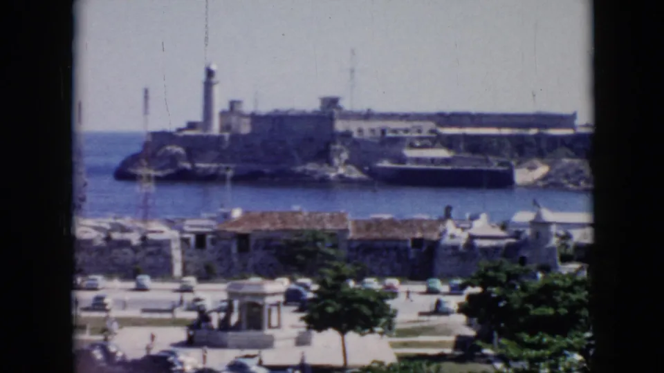 1956 Cuba 8mm frame showing a distant coastal fortress with a lighthouse across blue water, with stone waterfront buildings, trees, and cars in the foreground.