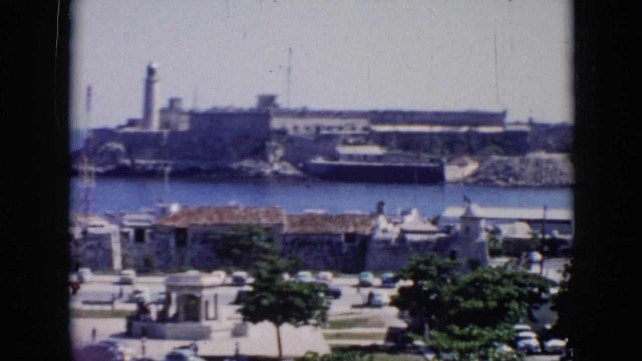 Harbor view with lighthouse, fortress walls, boat, waterfront roofs, trees, and street traffic.