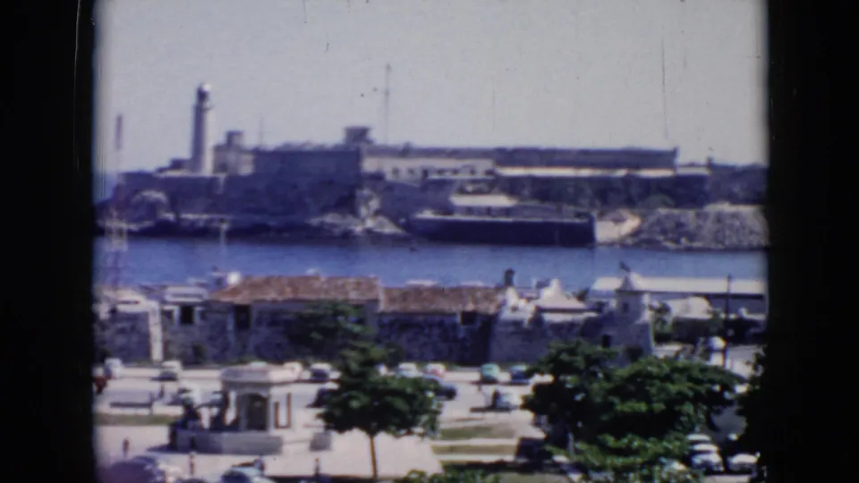 1956 Cuba 8mm frame with a harbor-facing view of a fortress and lighthouse, a dark boat near the shore, and low waterfront buildings below.